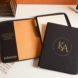 Könnte beinhalten: Schwarzes Leder-Portfolio mit goldenem Monogramm und den Initialen "KA". Das Portfolio ist geöffnet und zeigt einen Notizblock und einen Stift. Der Einband trägt den Namen "Johnson" in goldfarbenen Buchstaben und ein Zitat.