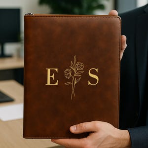 May include: A brown leather portfolio with a gold zipper, held by a person. The front features the gold letters "E" and "S" with a floral design between them. The portfolio is rectangular.