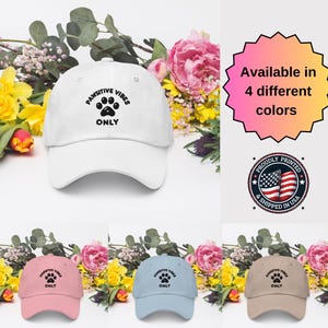 May include: White, pink, light blue, and brown baseball caps with a paw print and the text "Pawsitive Vibes Only" embroidered on the front. The caps are available in four different colors.