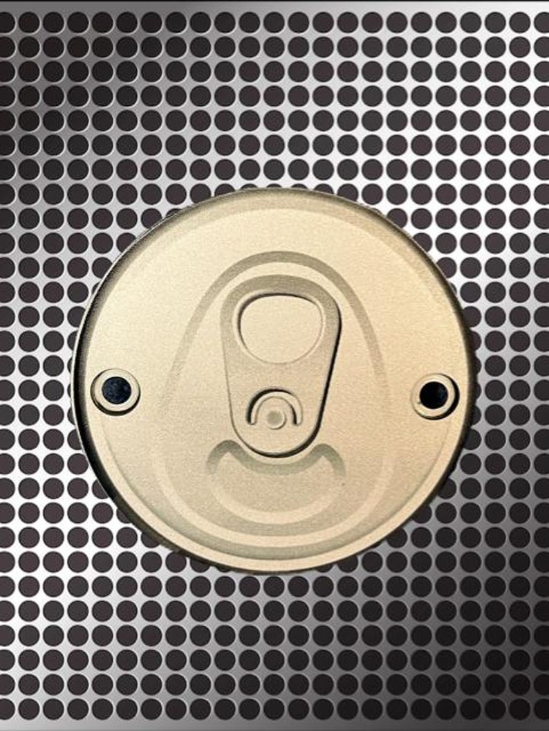 Pu&ograve; includere: Primo piano di un coperchio di lattina dorato con linguetta e due piccoli rivetti neri. Il coperchio &egrave; centrato su una superficie metallica grigia con un motivo di piccoli punti neri distanziati.