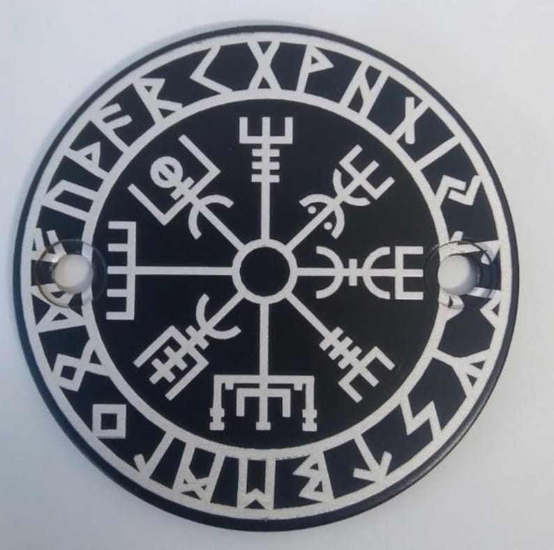 Pu&ograve; includere: Un emblema circolare in bianco e nero con il simbolo Vegv&iacute;sir, una bussola vichinga. Il design include iscrizioni runiche attorno al bordo e un design a bussola centrale. Due fori sono visibili sui lati dell'emblema.