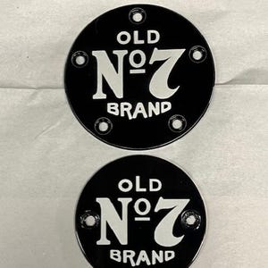 Pu&ograve; includere: Due targhe metalliche nere e circolari con la scritta "OLD N&deg;7 BRAND" in bianco. Ogni targa ha quattro fori equidistanti sul bordo. Le targhe sono su una superficie grigio chiaro.