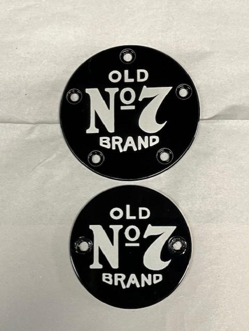 Pu&ograve; includere: Due targhe metalliche nere e circolari con la scritta "OLD N&deg;7 BRAND" in bianco. Ogni targa ha quattro fori equidistanti sul bordo. Le targhe sono su una superficie grigio chiaro.