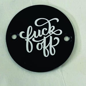 Pu&ograve; includere: Un oggetto circolare nero con la scritta bianca "fuck off" in corsivo. L'oggetto presenta due piccoli fori vicino ai bordi. Lo sfondo &egrave; di un colore chiaro e neutro.