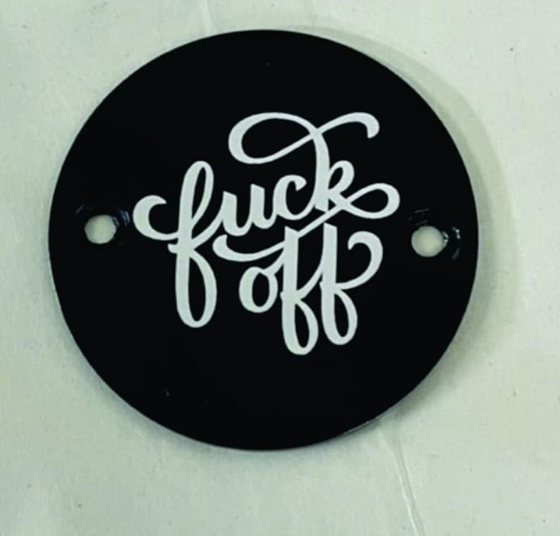 Pu&ograve; includere: Un oggetto circolare nero con la scritta bianca "fuck off" in corsivo. L'oggetto presenta due piccoli fori vicino ai bordi. Lo sfondo &egrave; di un colore chiaro e neutro.
