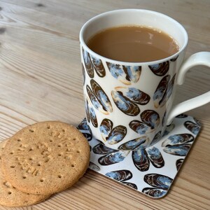 May include: A white ceramic mug filled with tea, featuring a pattern of blue and brown mussel shells. The mug sits on a matching coaster, next to two round biscuits. The mug has a white handle.