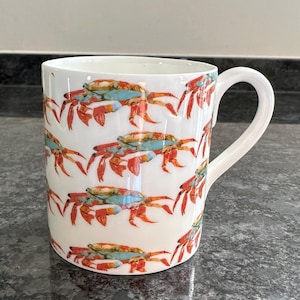 May include: White ceramic mug featuring a repeating pattern of crabs. The crabs have red claws and legs, blue and yellow shells, set against a white background. The mug has a curved handle.