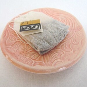 K&ouml;nnte beinhalten: Eine rosa Keramik-Teetasse-Untertasse mit Blumenmuster. Ein TAZO-Teebeutel liegt auf der Untertasse.