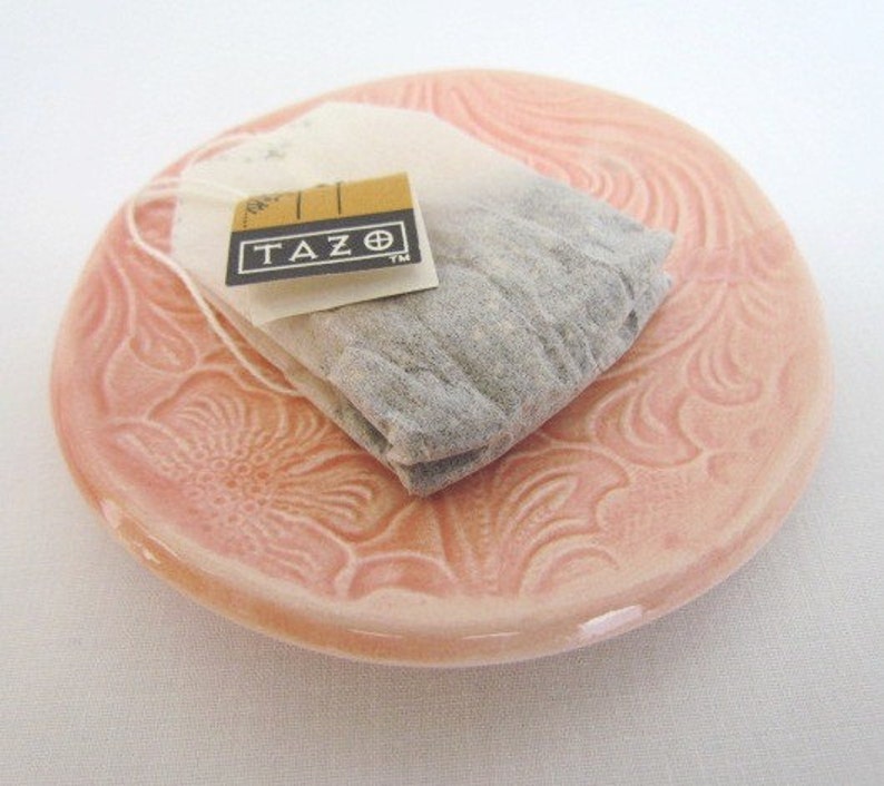 K&ouml;nnte beinhalten: Eine rosa Keramik-Teetasse-Untertasse mit Blumenmuster. Ein TAZO-Teebeutel liegt auf der Untertasse.