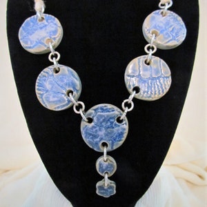Puede incluir: Un collar de cadena de plata con colgantes redondos de cerámica azul y blanca. Los colgantes tienen una superficie texturizada y están conectados por eslabones de cadena de plata.