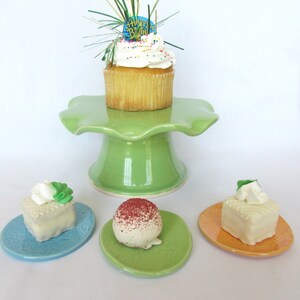 K&ouml;nnte beinhalten: Ein gr&uuml;ner Keramik-Kuchenst&auml;nder mit einem wei&szlig;en Cupcake mit Streuseln und einem "Happy Birthday"-Topper. Drei kleinere Teller mit einem wei&szlig;en Kuchenquadrat, einer roten und wei&szlig;en Kuchenkugel und einem wei&szlig;en Kuchenquadrat mit einem gr&uuml;nen Blatt obenauf.
