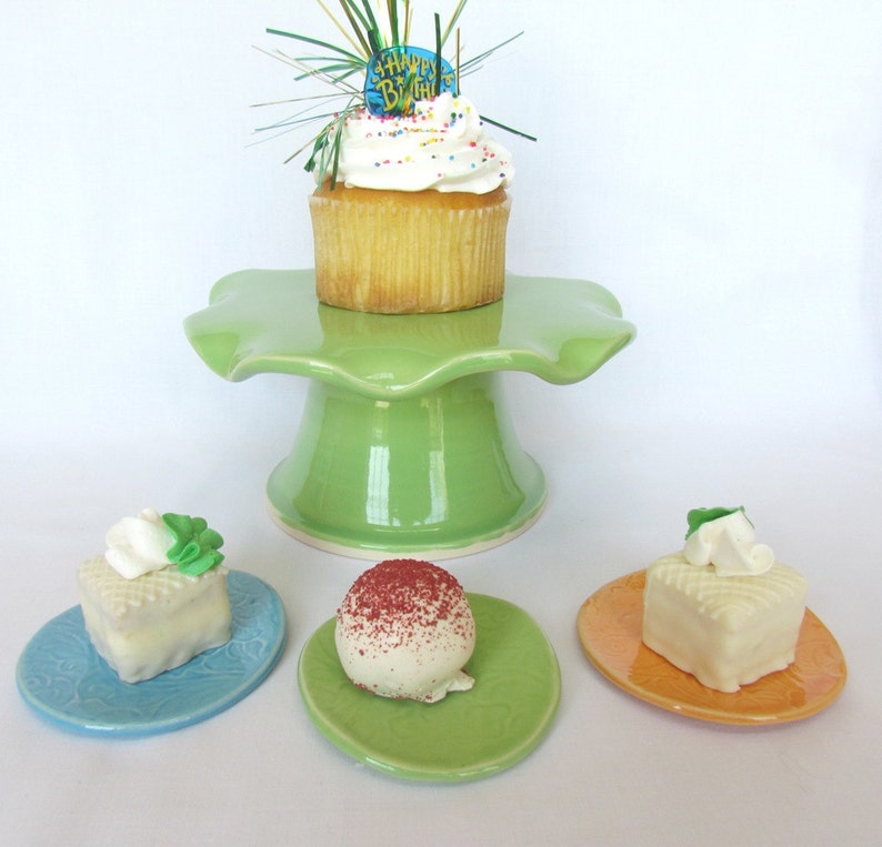 K&ouml;nnte beinhalten: Ein gr&uuml;ner Keramik-Kuchenst&auml;nder mit einem wei&szlig;en Cupcake mit Streuseln und einem "Happy Birthday"-Topper. Drei kleinere Teller mit einem wei&szlig;en Kuchenquadrat, einer roten und wei&szlig;en Kuchenkugel und einem wei&szlig;en Kuchenquadrat mit einem gr&uuml;nen Blatt obenauf.