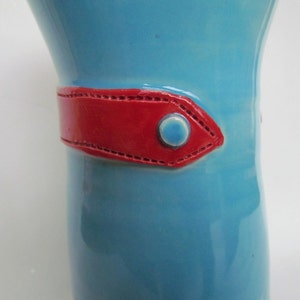 Może przedstawiać: Jasnoniebieski ceramiczny wazon z czerwonym paskiem ze skóry i niebieskim guzikiem.