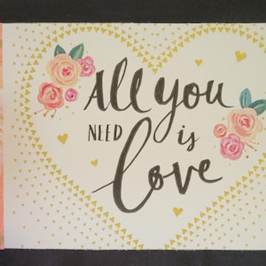 May include: A rectangular card with the phrase "All you need is Love" in black script. The card features a heart-shaped design with floral accents in pink and yellow, and a decorative pink and gold patterned edge.