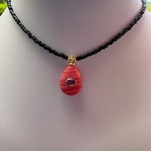 May include: A necklace featuring a strand of small black beads and a teardrop-shaped pendant. The pendant is coral-colored with horizontal white stripes and a central oval-shaped dark purple stone set in a gold-toned metal frame at the top.