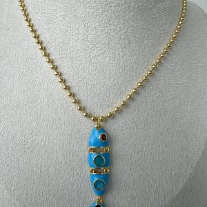Puede incluir: Collar dorado con un colgante en forma de pez. El pez es de color azul claro con detalles rojos, turquesa y azul oscuro. La cadena está hecha de pequeñas cuentas redondas.