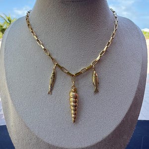 Puede incluir: Collar dorado con diseño de eslabones. El collar presenta tres colgantes: dos peces y una concha en espiral. La joya se exhibe en un maniquí con un fondo de cielo azul.