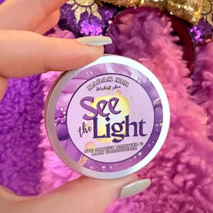 Könnte beinhalten: Eine silberne Dose mit einem lila und goldenen Etikett, auf dem "Madam Mim Wicked Skin See the Light Eye Makeup Remover & Lash Conditioner" steht.