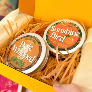 Puede incluir: Una caja de regalo amarilla contiene dos latas redondas de manteca corporal. Una lata dice "Dole Whipped" con "Pineapple & Vanilla Swirl Body Butter". La otra lata dice "Sunshine Bird" con "Freshly Squeezed Body Butter". La caja está llena de papel naranja.