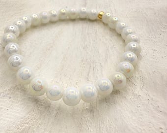 May include: A white beaded bracelet with iridescent sheen. The bracelet features round, pearl-like beads strung together with a gold-colored accent. The beads have a subtle, shimmering effect, reflecting light.