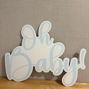 Op de afbeelding: Een wit houten bord met de woorden "Oh baby!" in lichtblauwe letters. Het bord is uitgesneden in de vorm van de woorden, met een lichtblauwe omtrek. Het bord staat op een houten oppervlak.