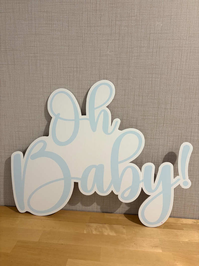 Op de afbeelding: Een wit houten bord met de woorden "Oh baby!" in lichtblauwe letters. Het bord is uitgesneden in de vorm van de woorden, met een lichtblauwe omtrek. Het bord staat op een houten oppervlak.
