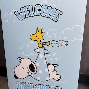 Op de afbeelding: Een lichtblauw babyborrelbord met Snoopy en Woodstock. Het bord heeft de woorden "WELCOME" en "BABY SHOWER" in blauwe letters. Woodstock houdt een banner vast met de tekst "IT'S A BOY!"
