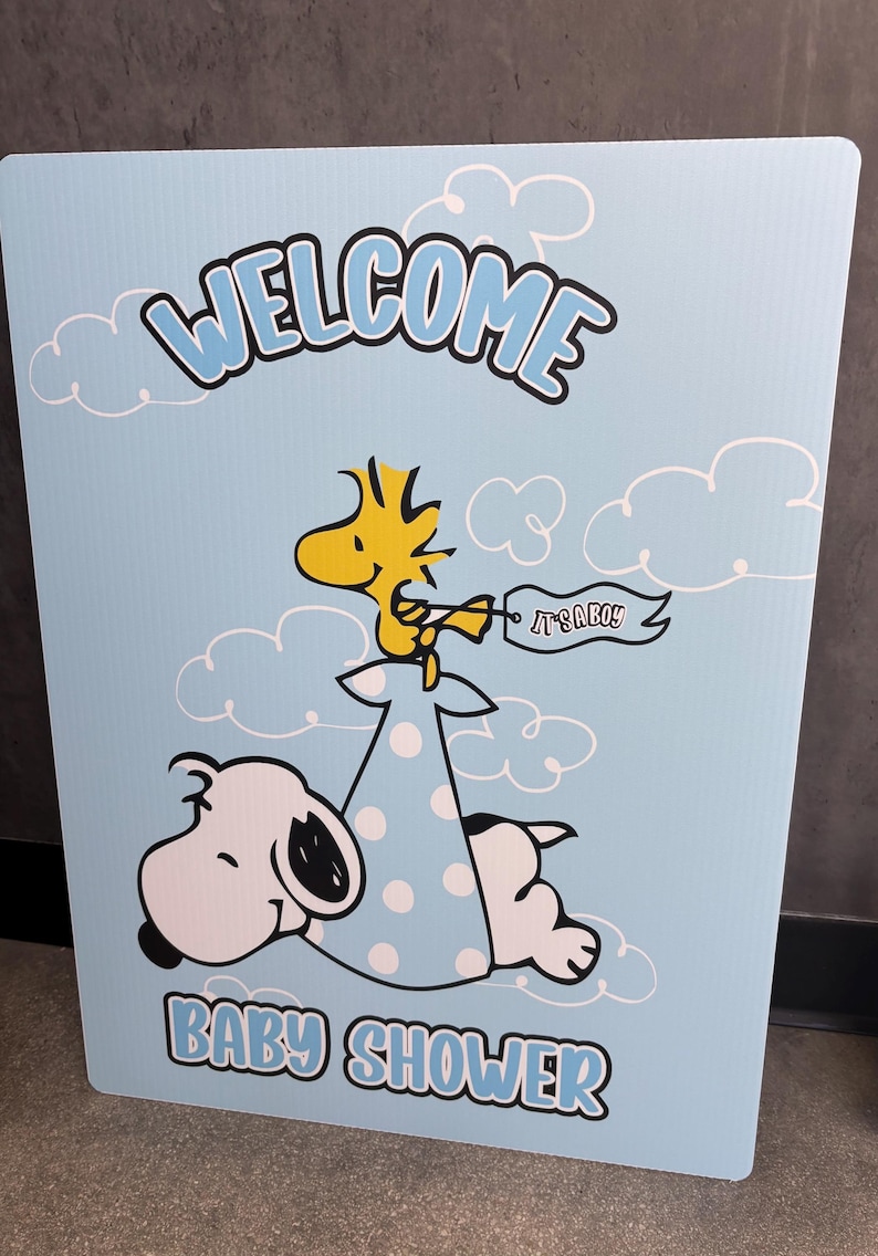 Op de afbeelding: Een lichtblauw babyborrelbord met Snoopy en Woodstock. Het bord heeft de woorden "WELCOME" en "BABY SHOWER" in blauwe letters. Woodstock houdt een banner vast met de tekst "IT'S A BOY!"