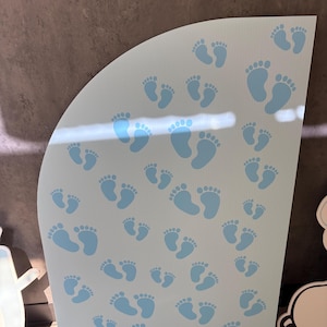 Op de afbeelding: Een lichtblauw, gebogen bord met talrijke babyvoetjes in een lichtere tint blauw. De voetafdrukken vari&euml;ren in grootte, wat verschillende leeftijden suggereert. Het bord is waarschijnlijk voor een babyshower of kinderkamerdecoratie.