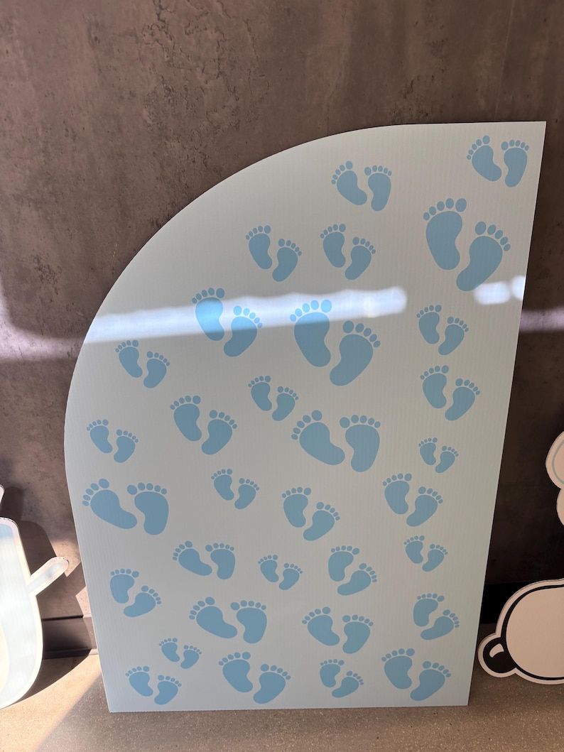Op de afbeelding: Een lichtblauw, gebogen bord met talrijke babyvoetjes in een lichtere tint blauw. De voetafdrukken vari&euml;ren in grootte, wat verschillende leeftijden suggereert. Het bord is waarschijnlijk voor een babyshower of kinderkamerdecoratie.