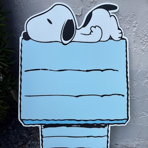 Op de afbeelding: Een Snoopy-tuinbord met Snoopy die slaapt op zijn hondenhok. Het hondenhok is lichtblauw met zwarte randen en horizontale lijnen. Het bord is gemaakt van duurzaam materiaal en is geschikt voor buitengebruik.