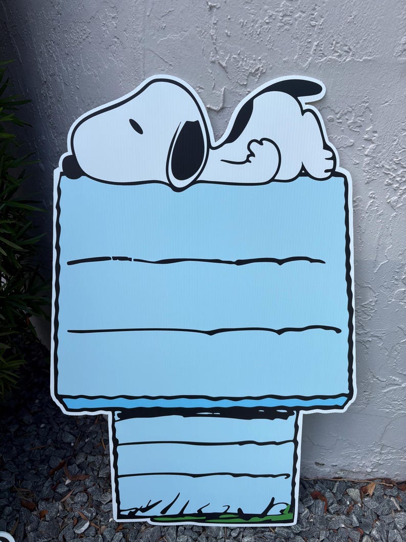 Op de afbeelding: Een Snoopy-tuinbord met Snoopy die slaapt op zijn hondenhok. Het hondenhok is lichtblauw met zwarte randen en horizontale lijnen. Het bord is gemaakt van duurzaam materiaal en is geschikt voor buitengebruik.