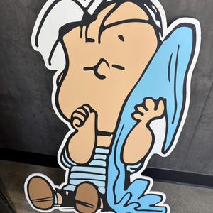 Op de afbeelding: Een grote cartoonuitsparing van een zittend personage met een bruine huid, gekleed in een blauw-wit gestreept shirt en bruine schoenen. Het personage houdt een lichtblauwe doek vast. De uitsparing heeft een witte omtrek.