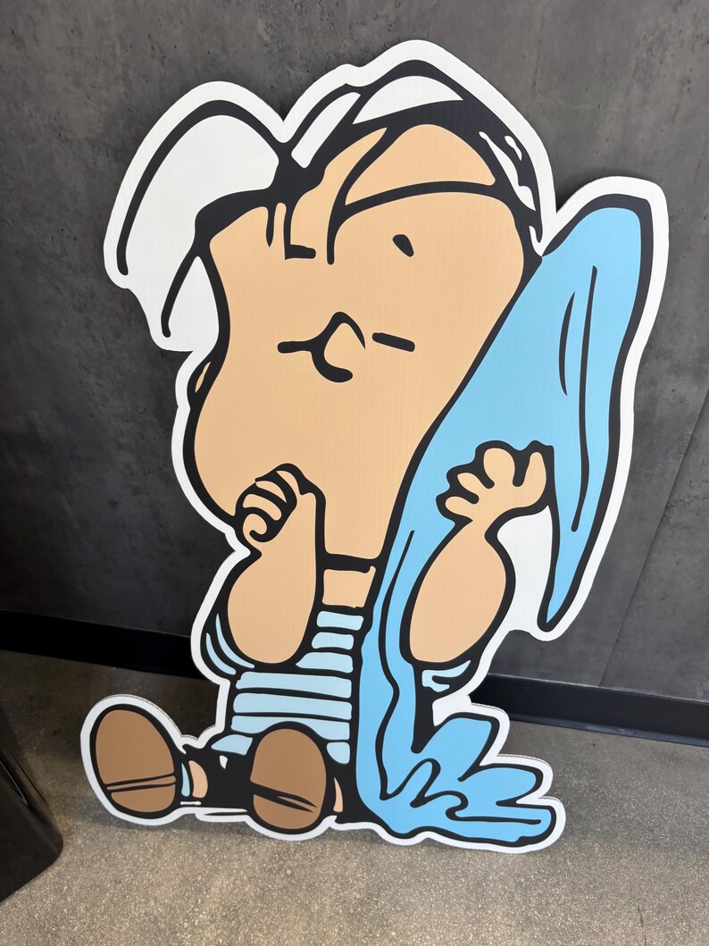 Op de afbeelding: Een grote cartoonuitsparing van een zittend personage met een bruine huid, gekleed in een blauw-wit gestreept shirt en bruine schoenen. Het personage houdt een lichtblauwe doek vast. De uitsparing heeft een witte omtrek.