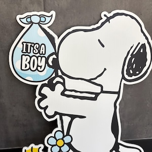 Op de afbeelding: Een witte Snoopy-uitsparing met een blauwe babybundel met de tekst "IT'S A BOY". Woodstock staat naast Snoopy en een blauwe bloem met een geel centrum is in de sc&egrave;ne. De uitsparing heeft een zwarte omtrek.