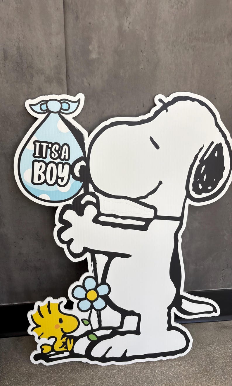 Op de afbeelding: Een witte Snoopy-uitsparing met een blauwe babybundel met de tekst "IT'S A BOY". Woodstock staat naast Snoopy en een blauwe bloem met een geel centrum is in de sc&egrave;ne. De uitsparing heeft een zwarte omtrek.