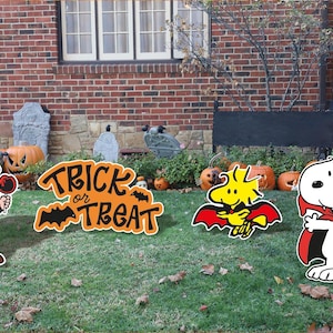 May include: Halloween yard decorations featuring Charlie Brown in a skeleton costume, Snoopy in a cape, and Woodstock in a bat costume. An orange sign reads "Trick or Treat" with bats. Pumpkins and a skeleton are also visible.
