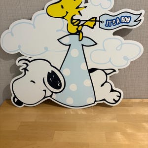 Op de afbeelding: Een grote, uitgesneden decoratie met Snoopy en Woodstock uit de Peanuts-strip. Woodstock houdt een banner vast met de tekst "IT'S A BOY". De figuren staan tegen een achtergrond van witte wolken en een lichtblauwe jurk met stippen.