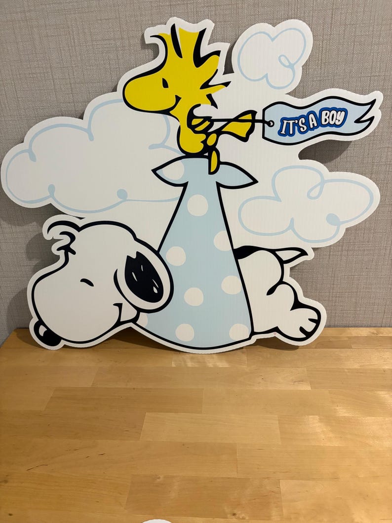 Op de afbeelding: Een grote, uitgesneden decoratie met Snoopy en Woodstock uit de Peanuts-strip. Woodstock houdt een banner vast met de tekst "IT'S A BOY". De figuren staan tegen een achtergrond van witte wolken en een lichtblauwe jurk met stippen.