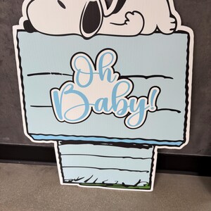 Op de afbeelding: Een lichtblauw bord met de tekst "Oh Baby!" in cursief, met een cartoon Snoopy-personage dat erop ligt. Het bord heeft een zwarte rand en is ontworpen voor een babyshower of gender reveal.