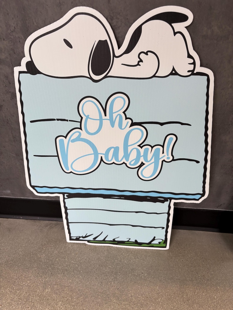 Op de afbeelding: Een lichtblauw bord met de tekst "Oh Baby!" in cursief, met een cartoon Snoopy-personage dat erop ligt. Het bord heeft een zwarte rand en is ontworpen voor een babyshower of gender reveal.