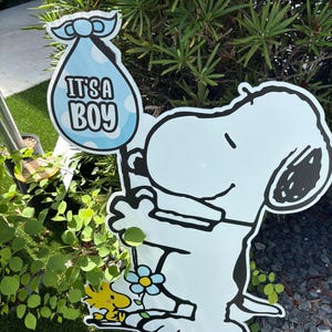 Op de afbeelding: Een lichtblauw Snoopy-tuinbord met een babybundel met de tekst "IT'S A BOY". Het bord toont ook Woodstock en een blauwe bloem. Het bord staat voor groen gebladerte.