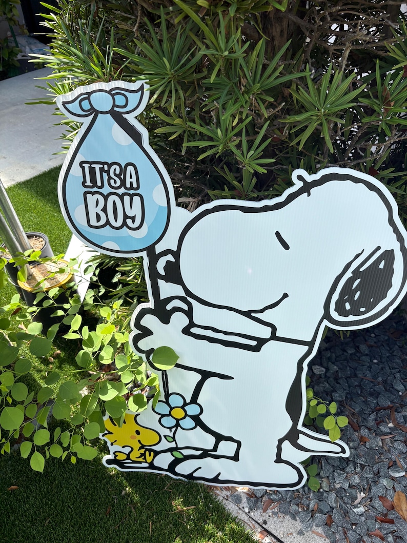 Op de afbeelding: Een lichtblauw Snoopy-tuinbord met een babybundel met de tekst "IT'S A BOY". Het bord toont ook Woodstock en een blauwe bloem. Het bord staat voor groen gebladerte.