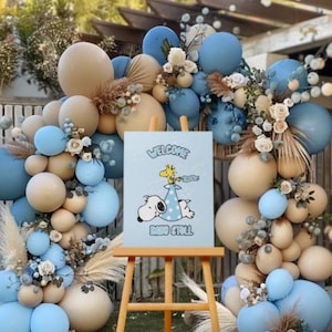 Op de afbeelding: Een babyborrel decoratie met een blauwe en beige ballonnenboog en bloemstukken. Een kleine houten ezel toont een bord met Snoopy en Woodstock, en de woorden "Welcome Baby Stoll."