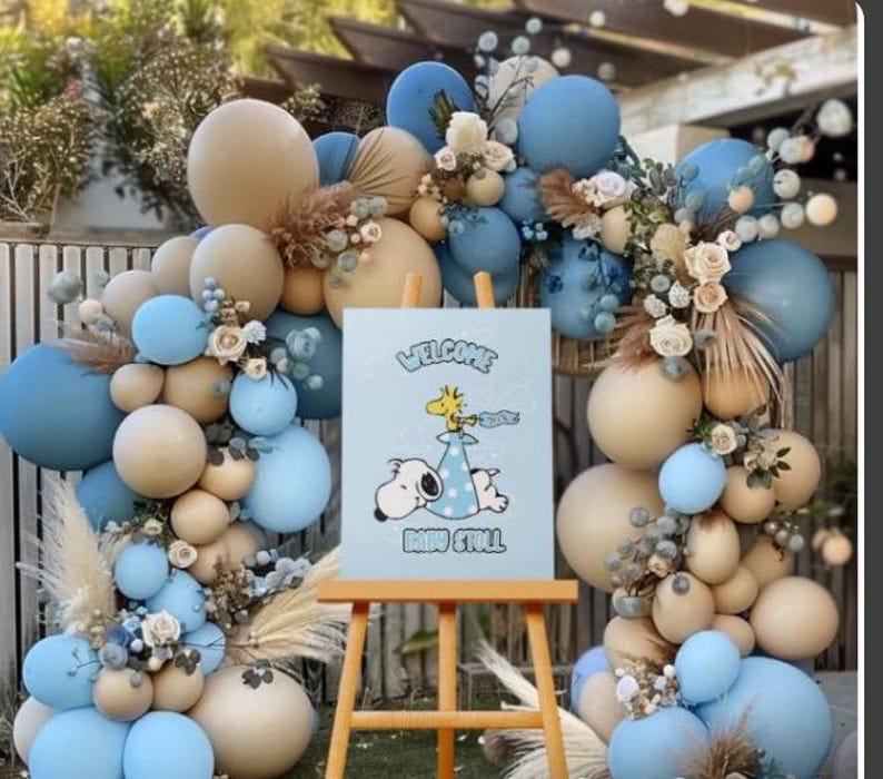 Op de afbeelding: Een babyborrel decoratie met een blauwe en beige ballonnenboog en bloemstukken. Een kleine houten ezel toont een bord met Snoopy en Woodstock, en de woorden "Welcome Baby Stoll."