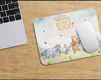 May include: A rectangular mousepad featuring a watercolor illustration of Winnie the Pooh and friends. The design includes Pooh, Piglet, Eeyore, and bees. A white computer mouse rests on the pad, next to a laptop on a wooden surface.