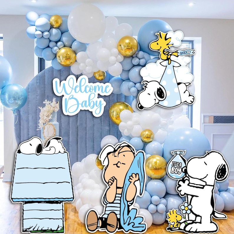 Op de afbeelding: Een babyborrel decoratie met een blauw-witte ballonnenboog met gouden accenten. Uitsparingen van Snoopy en Woodstock personages worden weergegeven, samen met de tekst "Welcome Baby" en "It's a Boy".