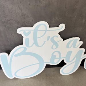 Op de afbeelding: Wit bord met de tekst "it's a boy" in lichtblauwe letters. Het bord heeft een decoratieve, afgeronde vorm en is geschikt voor een babyshower of gender reveal party.