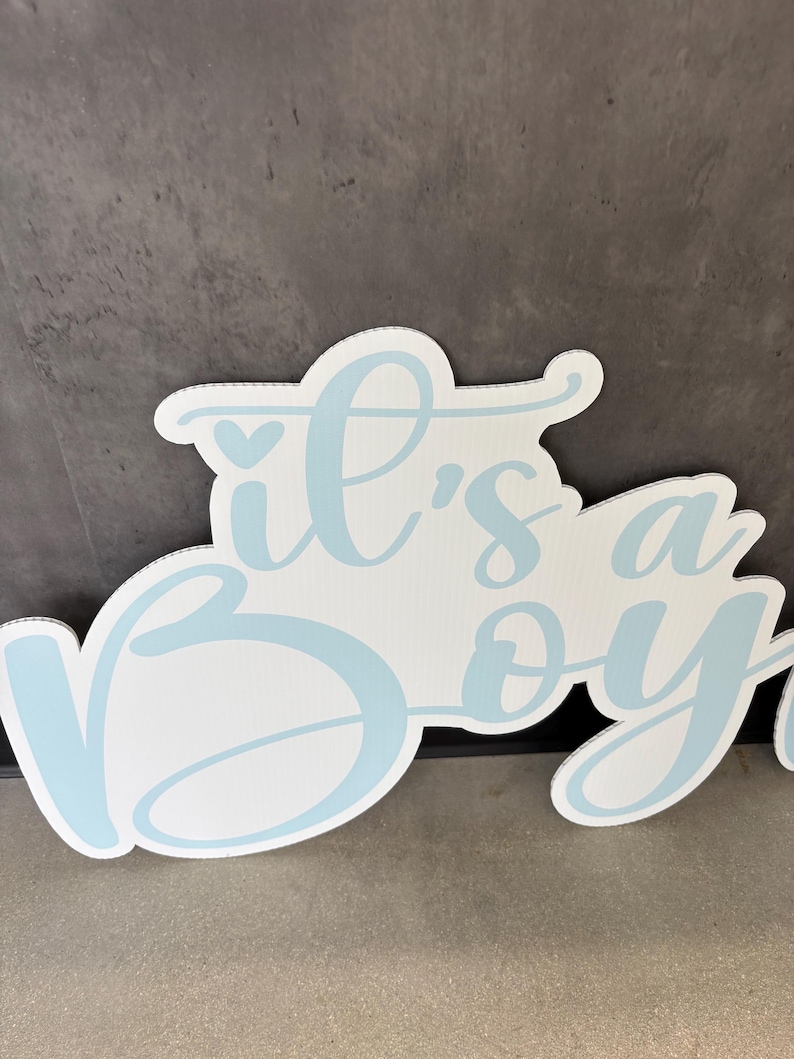 Op de afbeelding: Wit bord met de tekst "it's a boy" in lichtblauwe letters. Het bord heeft een decoratieve, afgeronde vorm en is geschikt voor een babyshower of gender reveal party.