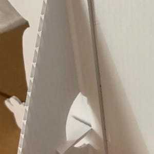 Op de afbeelding: Een witte golfkartonnen ezelrugsteun, ontworpen om rechtop te staan. De steun heeft een driehoekige vorm met een ingebouwde standaard voor stabiliteit. De randen hebben een geribbelde textuur.